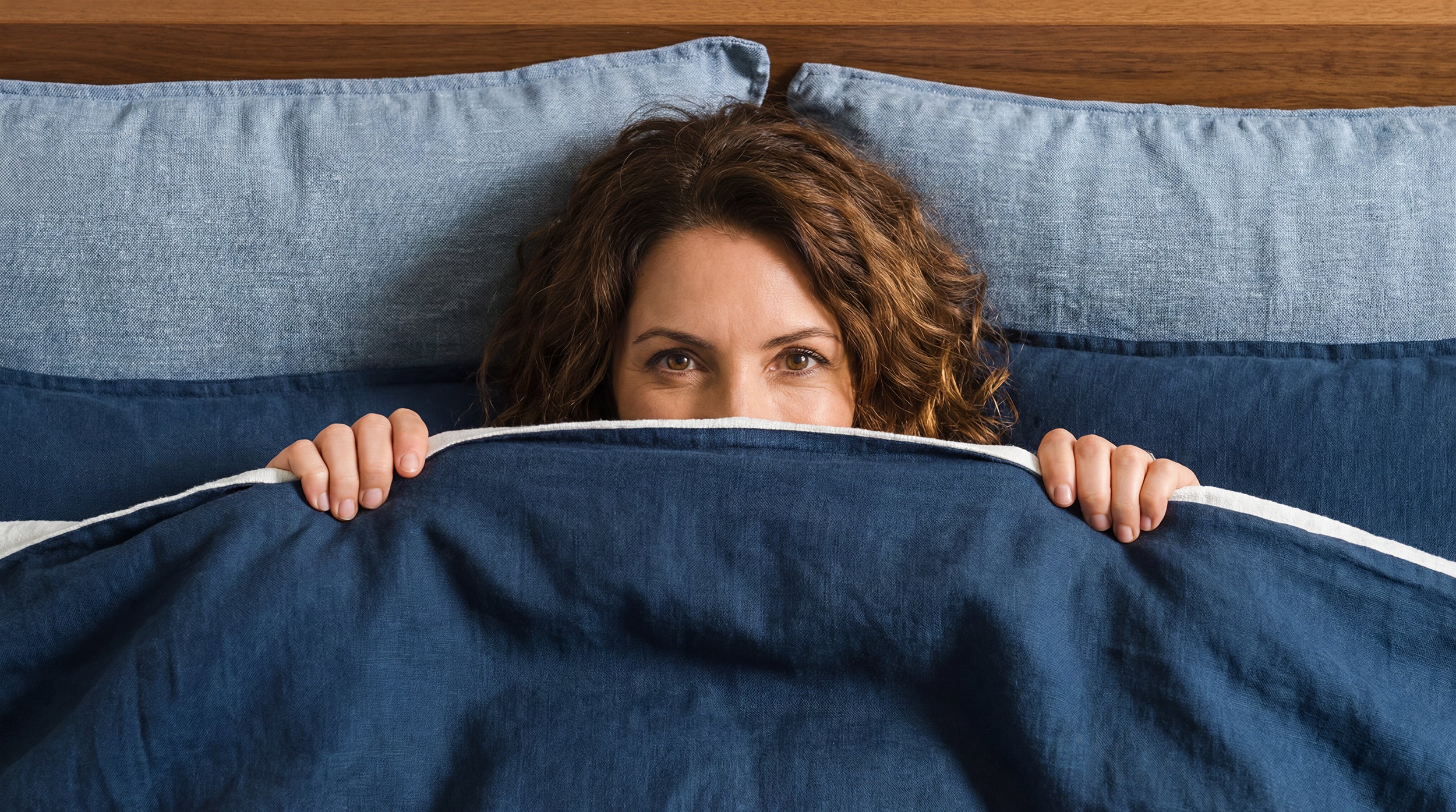 Woman peeking from under blue bedding on a bed with wooden headboard