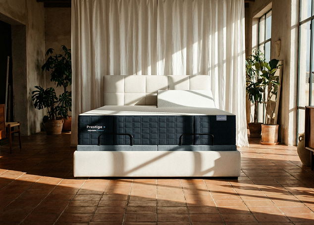 Modern bed with 'Prestige' branding in a sunlit room with plants and curtains.